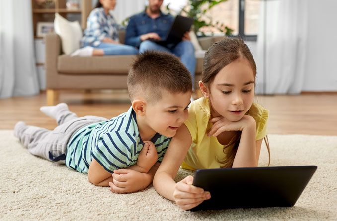 Menino e menina deitados no tapete mexendo no tablet.
